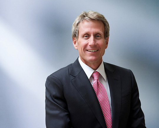 A man, Michael Sanders, with short, dark brown hair, wearing a black blazer, is smiling at the camera, standing against a blurred blue and white background."