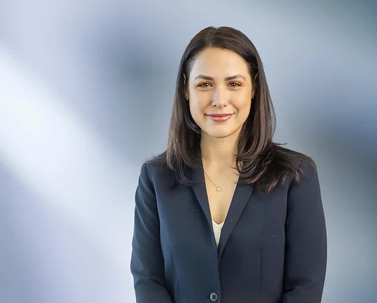 Woman, Alicia Birach, in a dark navy blazer standing against a softly blurred blue and white background, smiling confidently at the camera