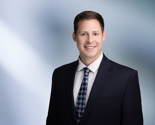 A man, Michael Cassar, with short, dark brown hair and a beard, wearing a black blazer and white shirt, is smiling at the camera, standing against a blurred blue and white background.”