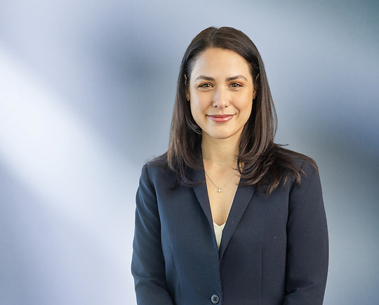 Woman, Alicia Birach, in a dark navy blazer standing against a softly blurred blue and white background, smiling confidently at the camera